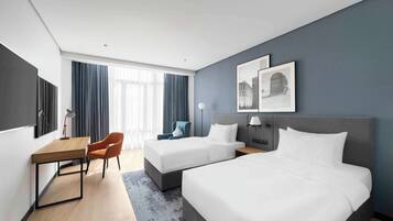 A hotel room with two beds, a desk, a chair, and a large window with curtains.