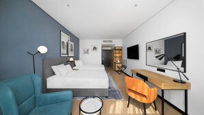 A modern hotel room with a bed, a desk, a chair, a TV, and artwork on the walls.