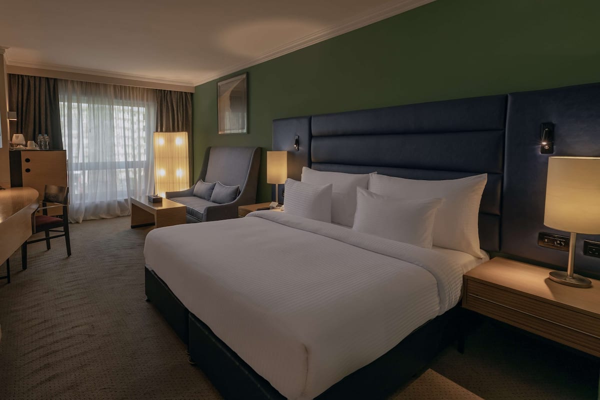 A hotel room with a large bed, a desk, a chair, and a nightstand with a lamp.