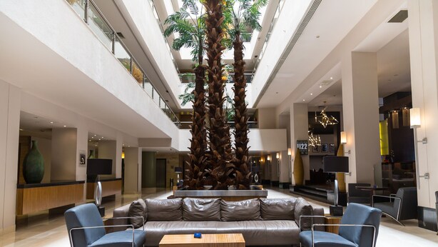 Um lobby moderno com área de estar central, uma grande árvore decorativa e teto alto com iluminação natural.