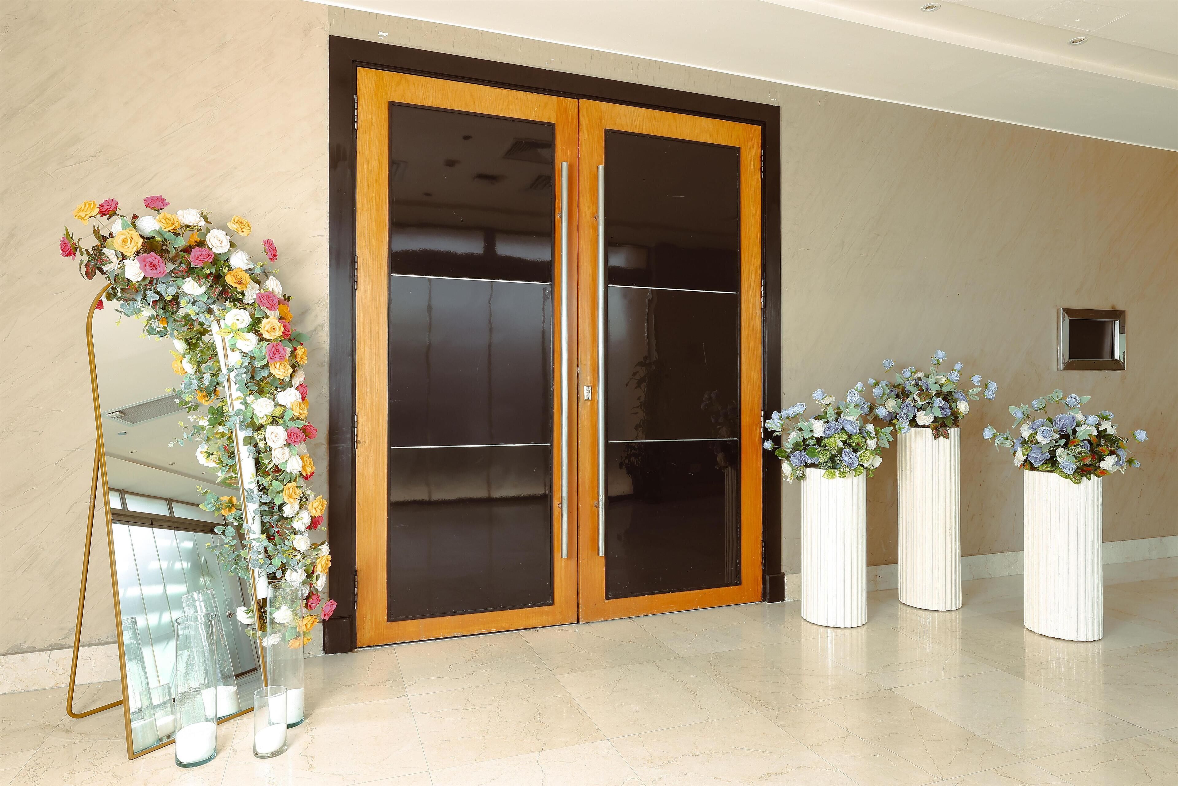A hallway with a large wooden door, a mirror, and three white vases with flowers.