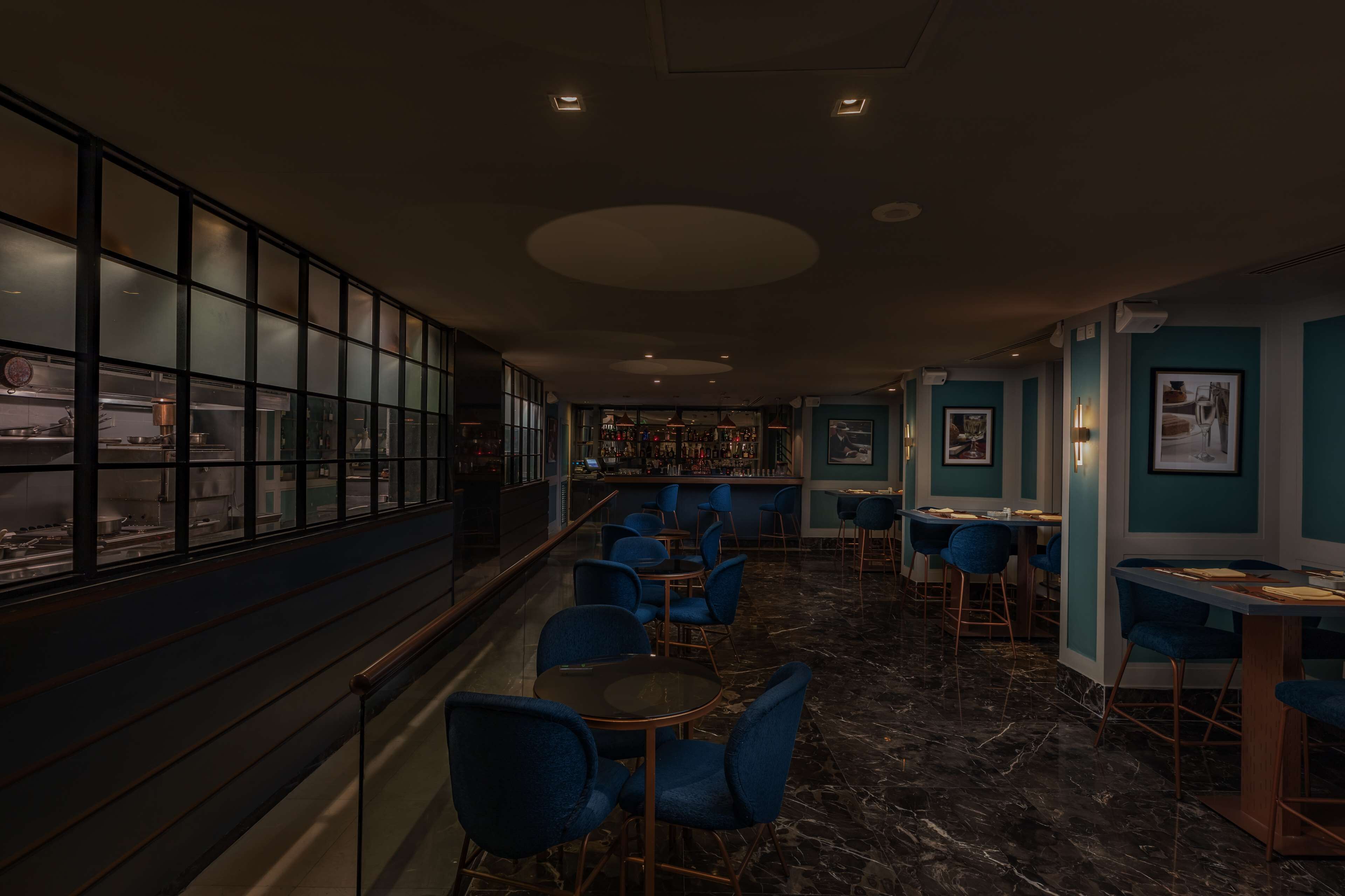 Zona de bar con poca luz, con sillas y mesas azules, una barra y vistas de la cocina a través de un vidrio satinado.