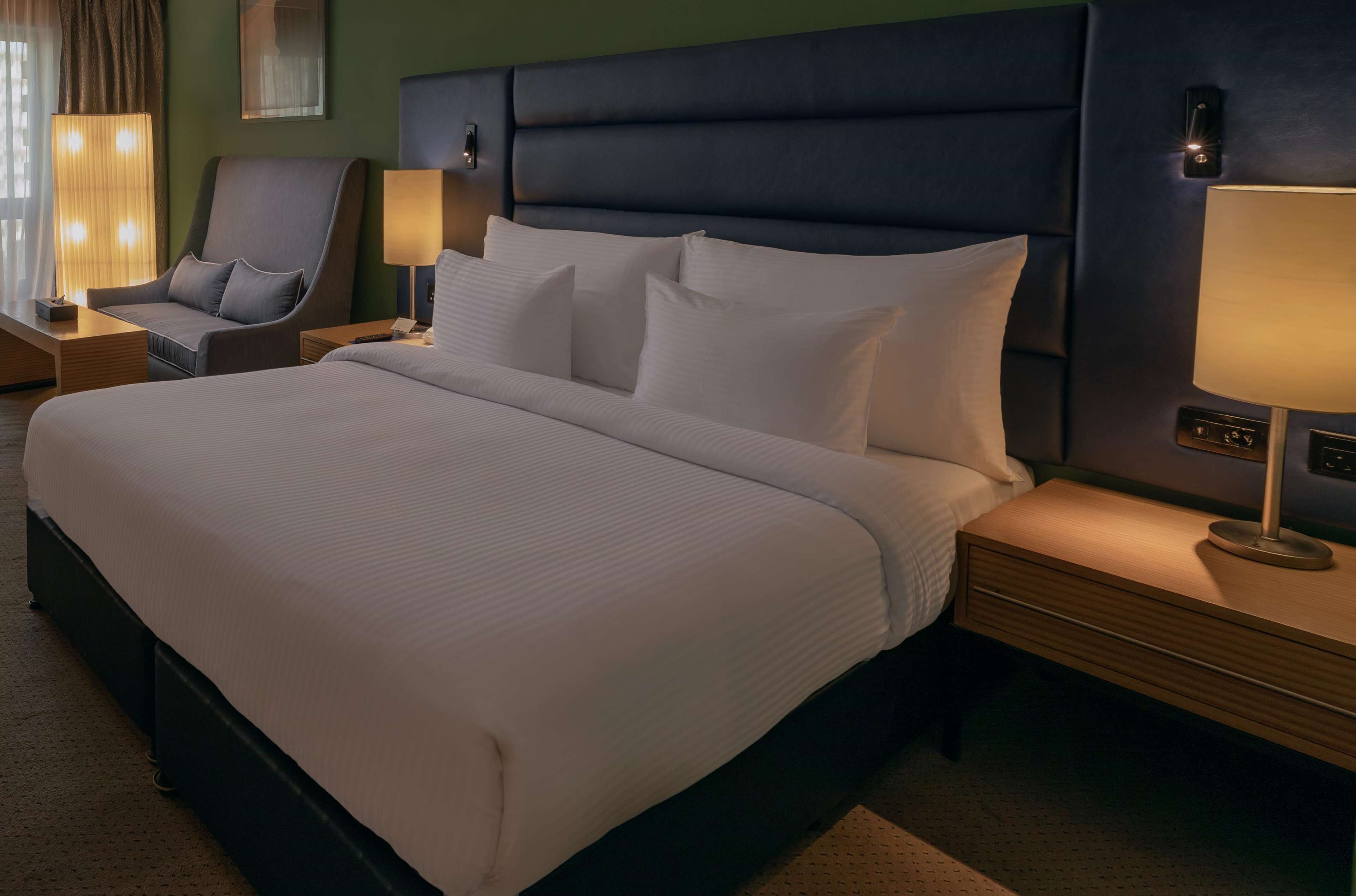 A hotel room with a large bed, a chair, two bedside tables with lamps, and a wall-mounted control panel.