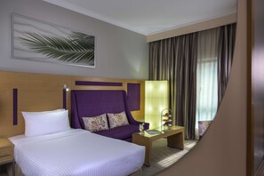Habitación de hotel con una cama grande, cabecera morada, mesita de noche, mesa auxiliar con lámpara, una obra de arte enmarcada en la pared y una ventana con cortinas.