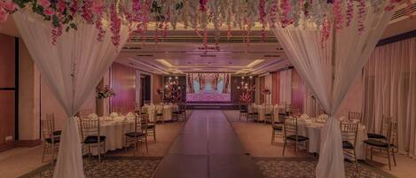 A banquet hall with a long table set for a formal event, draped with white fabric and pink flowers.