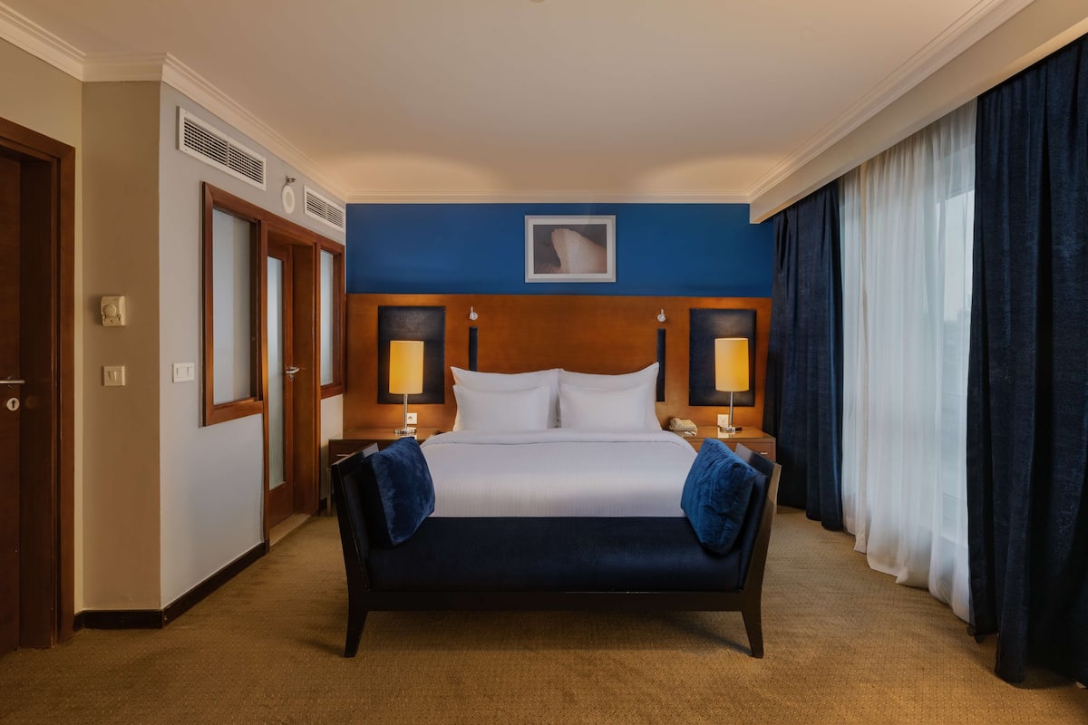 A hotel room with a large bed, two bedside tables with lamps, a headboard with a framed picture, and a large window with curtains.