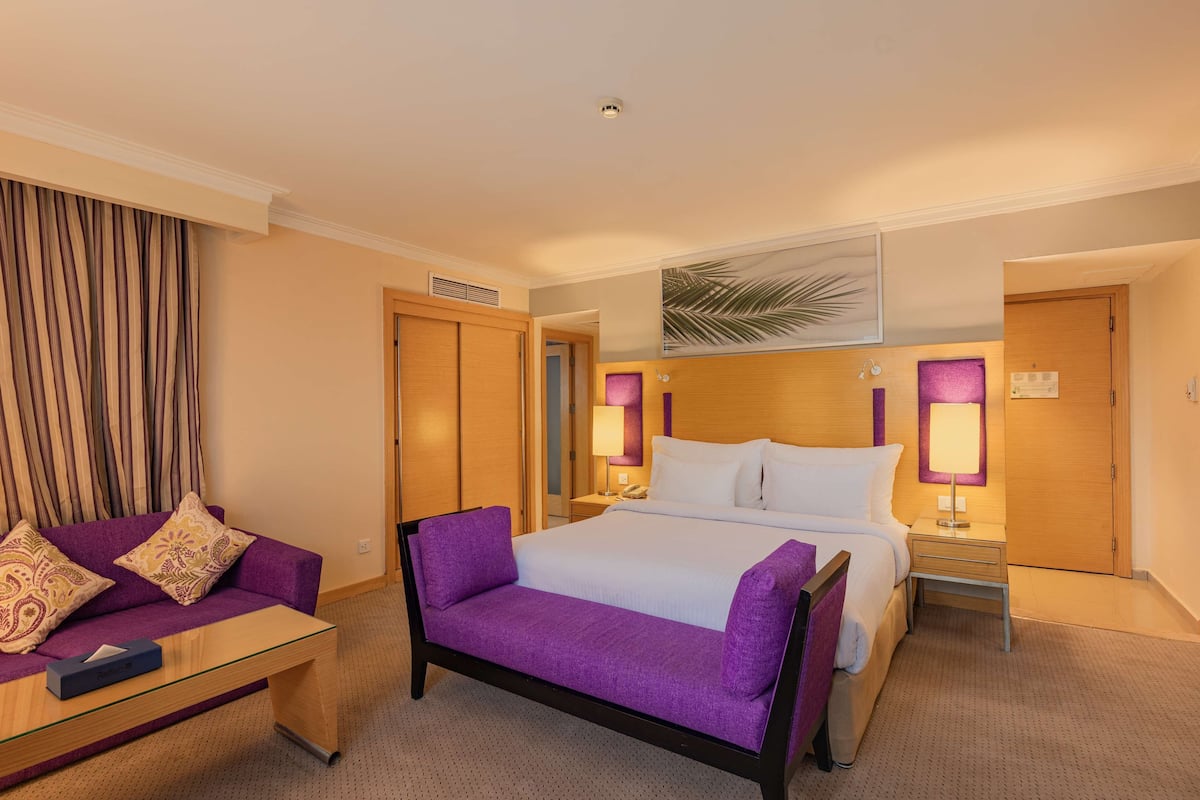 A hotel room with a bed, a purple sofa, a small table, and two bedside lamps.