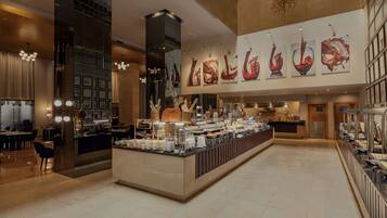 A modern hotel lobby with a central buffet area, a seating area with tables and chairs, and a large wall with abstract art.