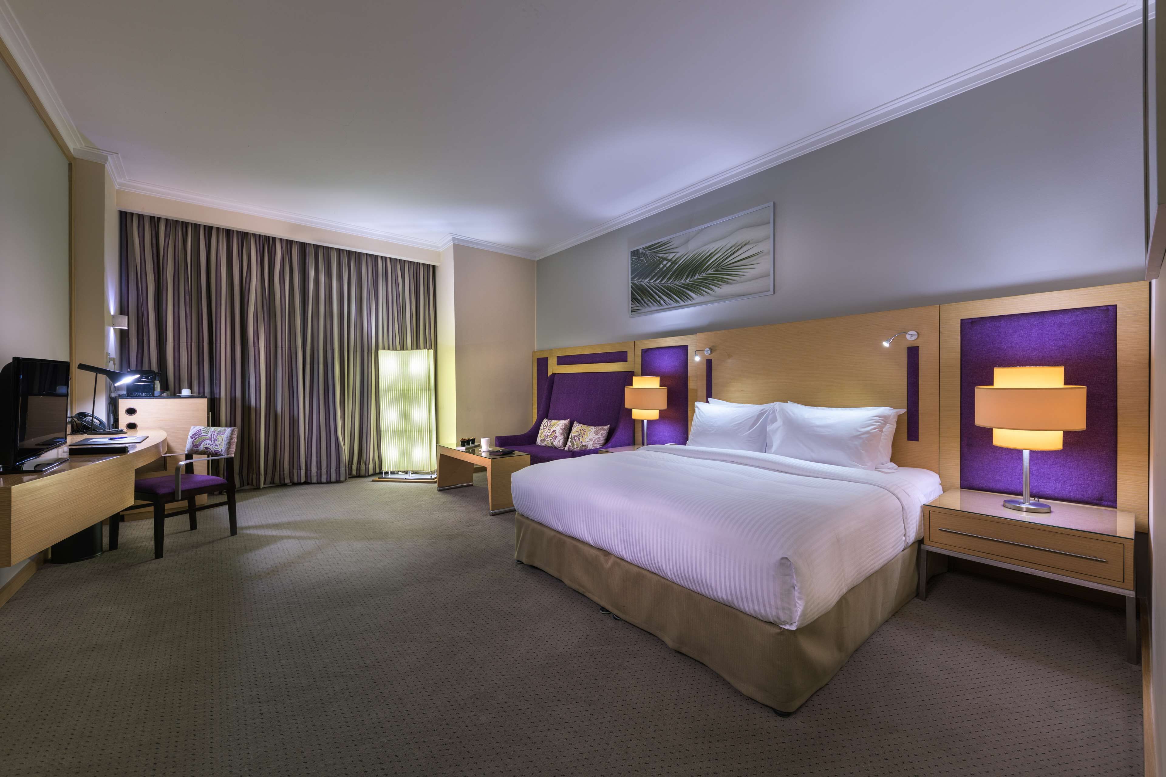 A hotel room with a large bed, a desk with a computer, a chair, a nightstand with a lamp, and a window with curtains.