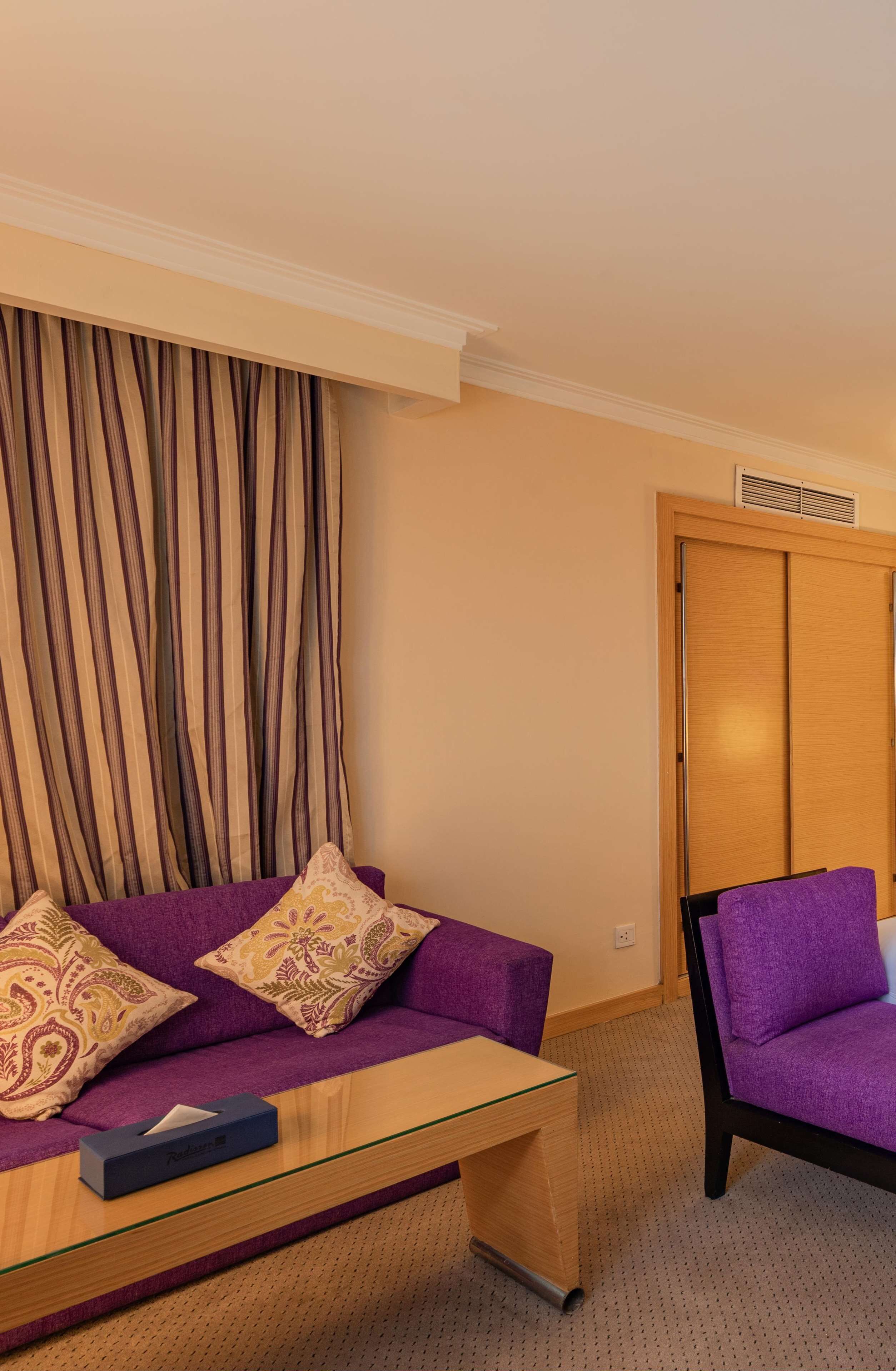 A room with a purple sofa, a glass coffee table, a purple armchair, and a wooden wardrobe.