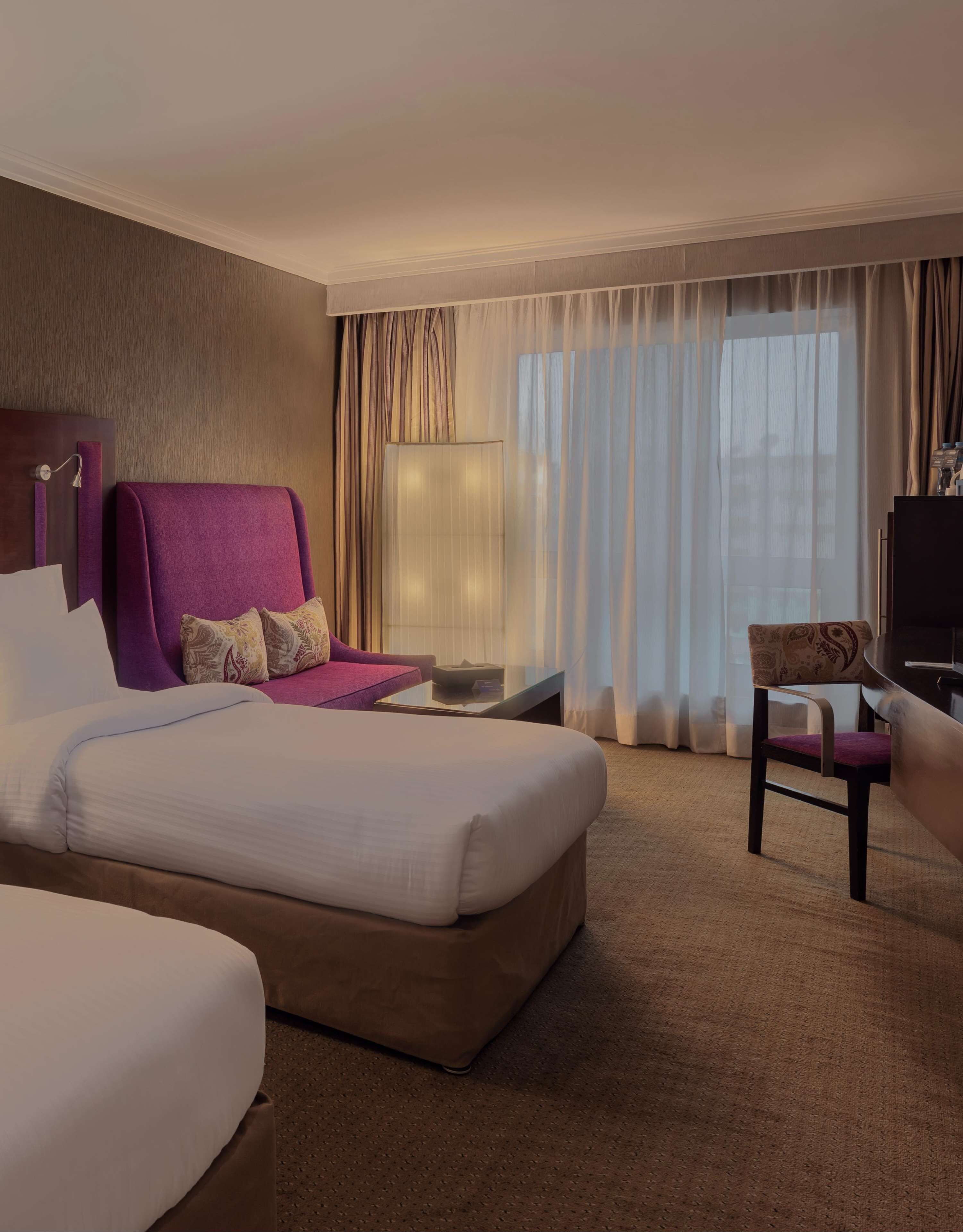 A hotel room with a large bed, a purple armchair, a small table, a TV, and a window with curtains.