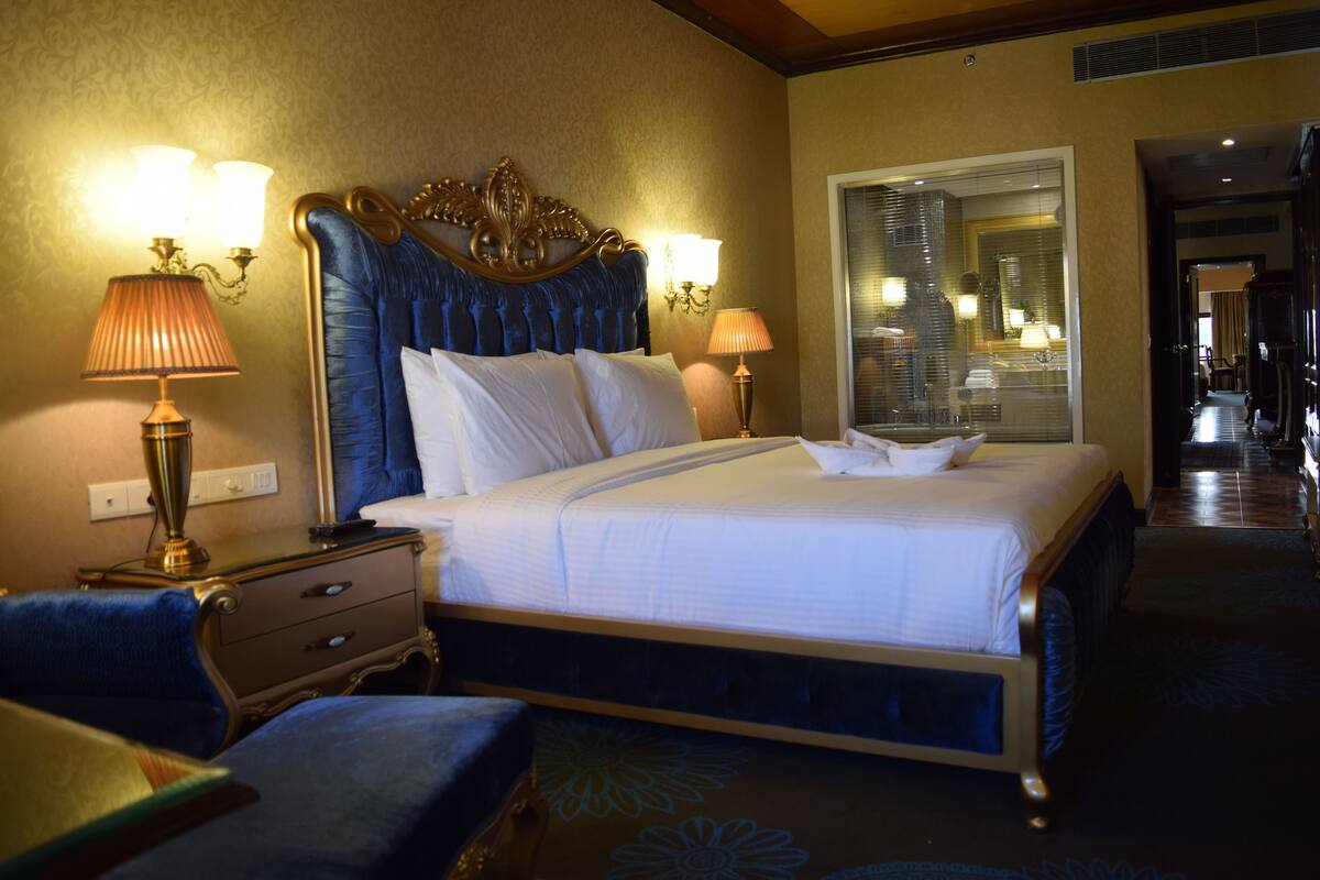 Quarto de hotel com uma cama grande, duas luminárias de cabeceira, uma mesinha de cabeceira, uma cadeira e um espelho.