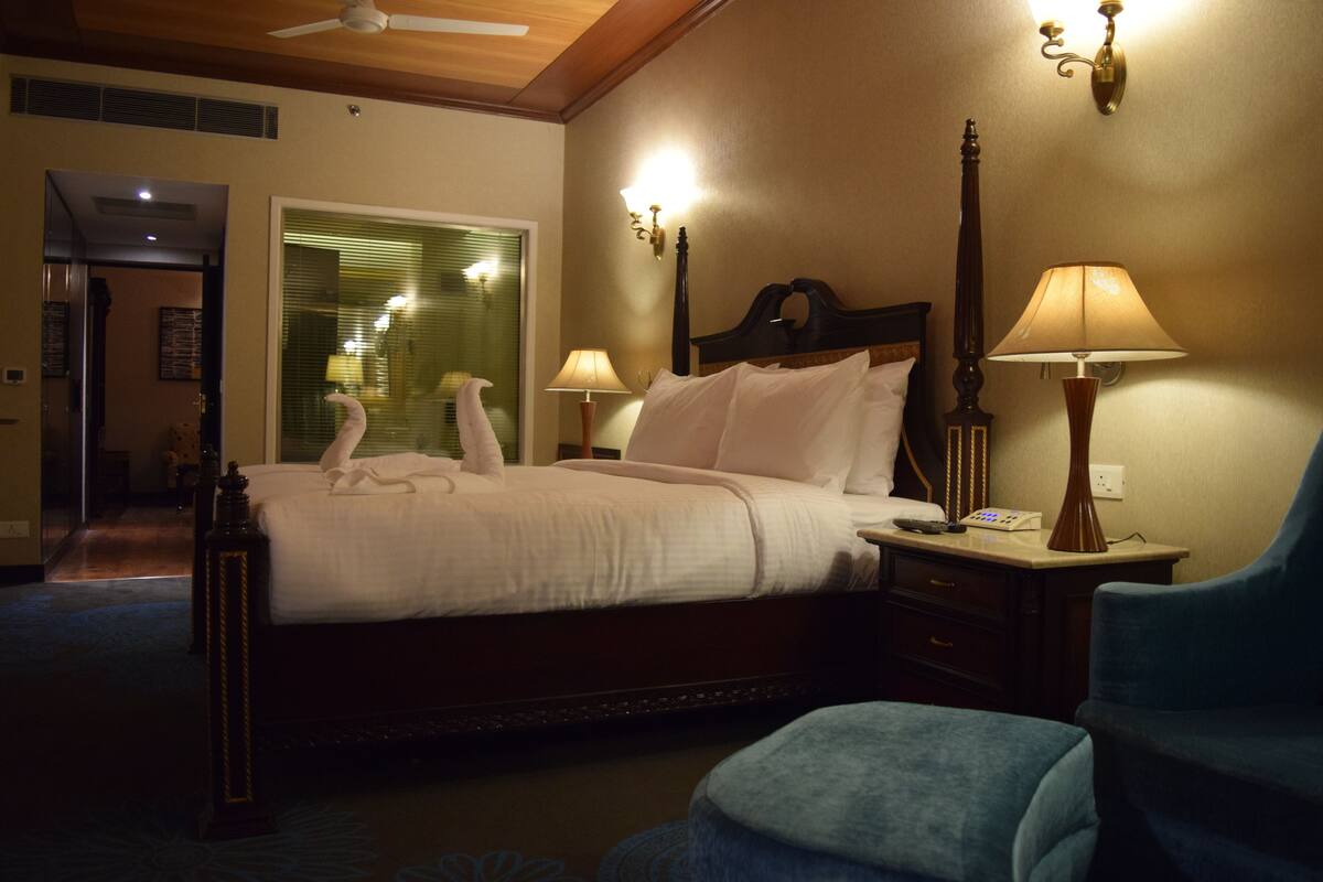 Quarto de hotel com uma cama grande, duas luminárias de cabeceira, um criado-mudo, uma cadeira e um ventilador de teto.