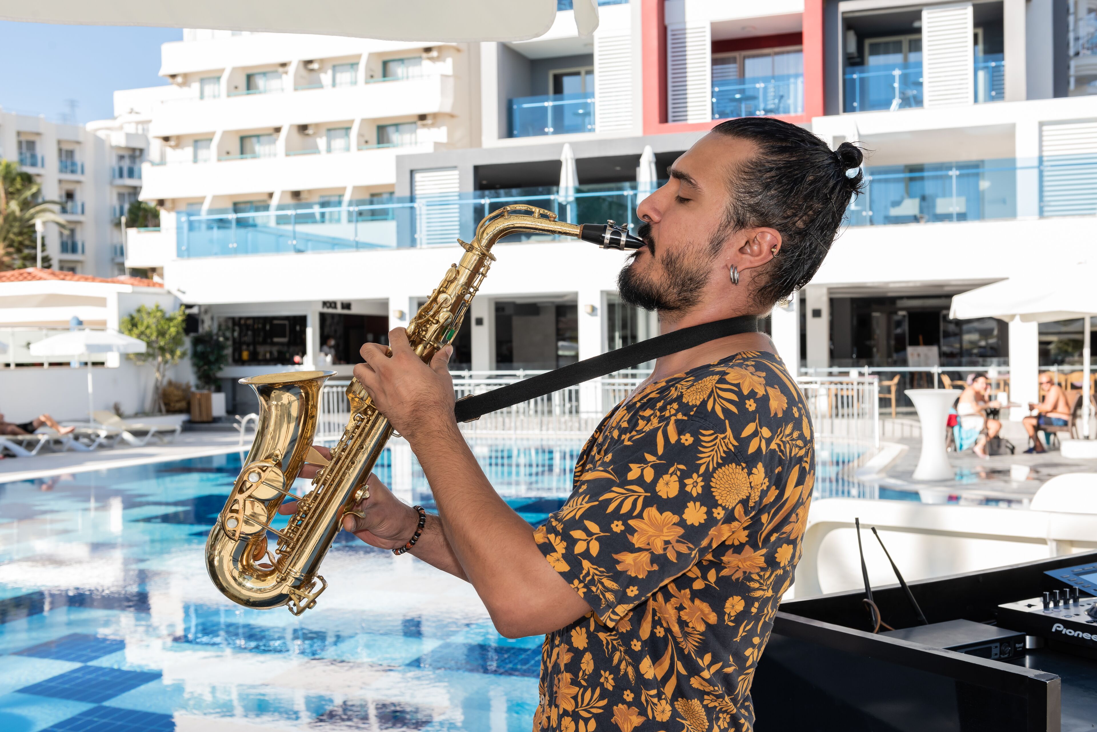 Un hombre tocando el saxofón junto a una piscina, con un edificio moderno al fondo.