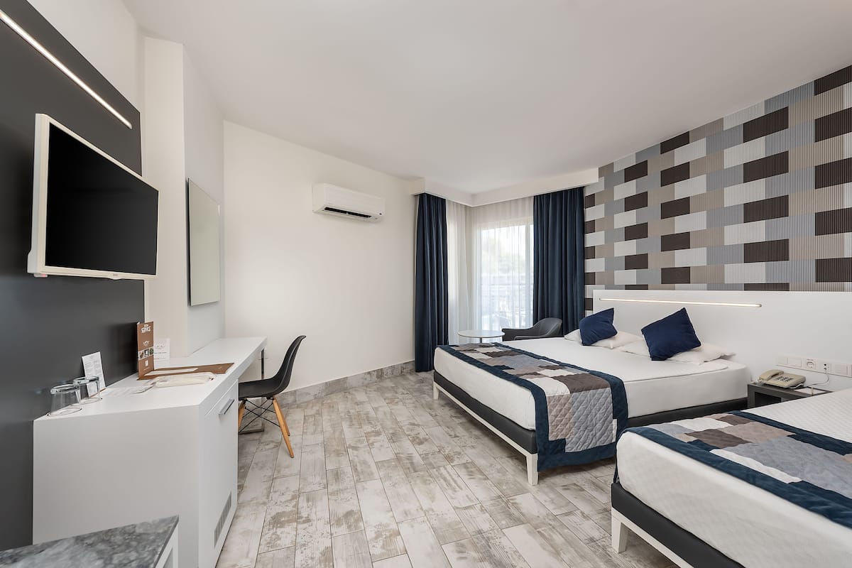 A modern hotel room with a desk, two beds, a flat-screen TV, and a large window with curtains.