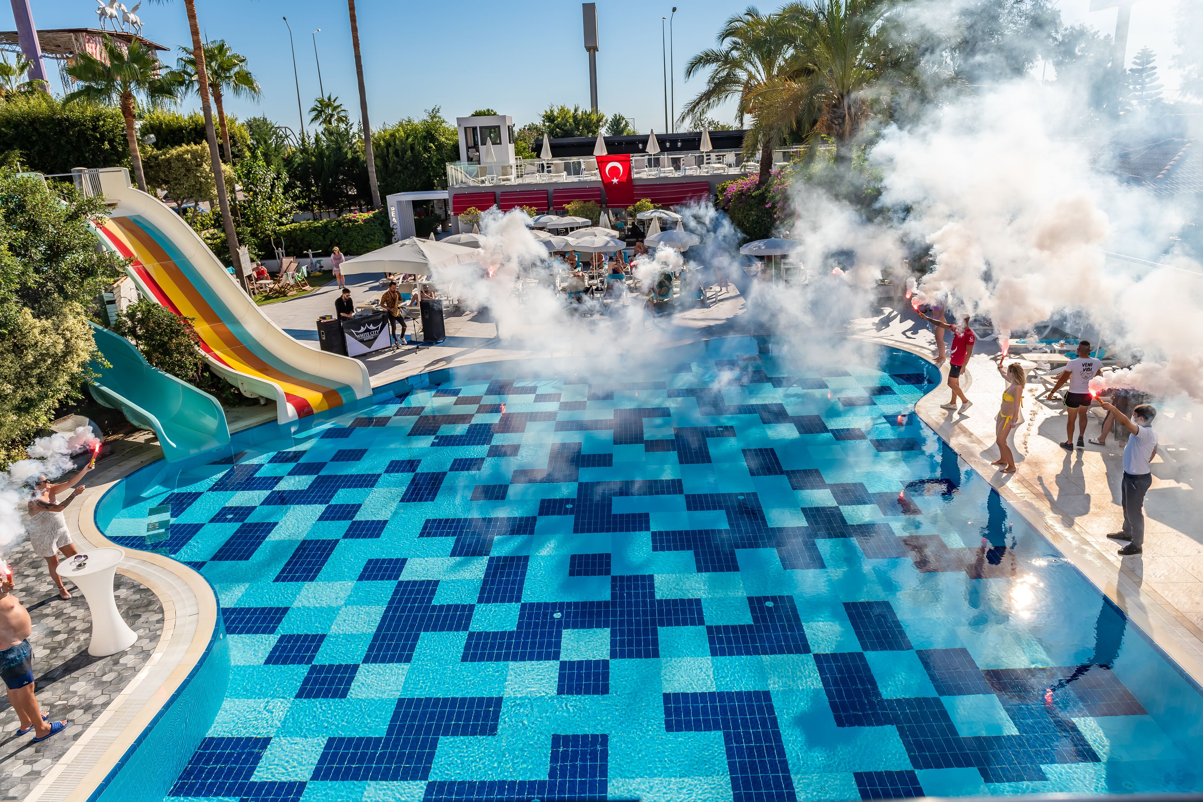 Una piscina con tobogán, rodeada de palmeras y una bandera roja.