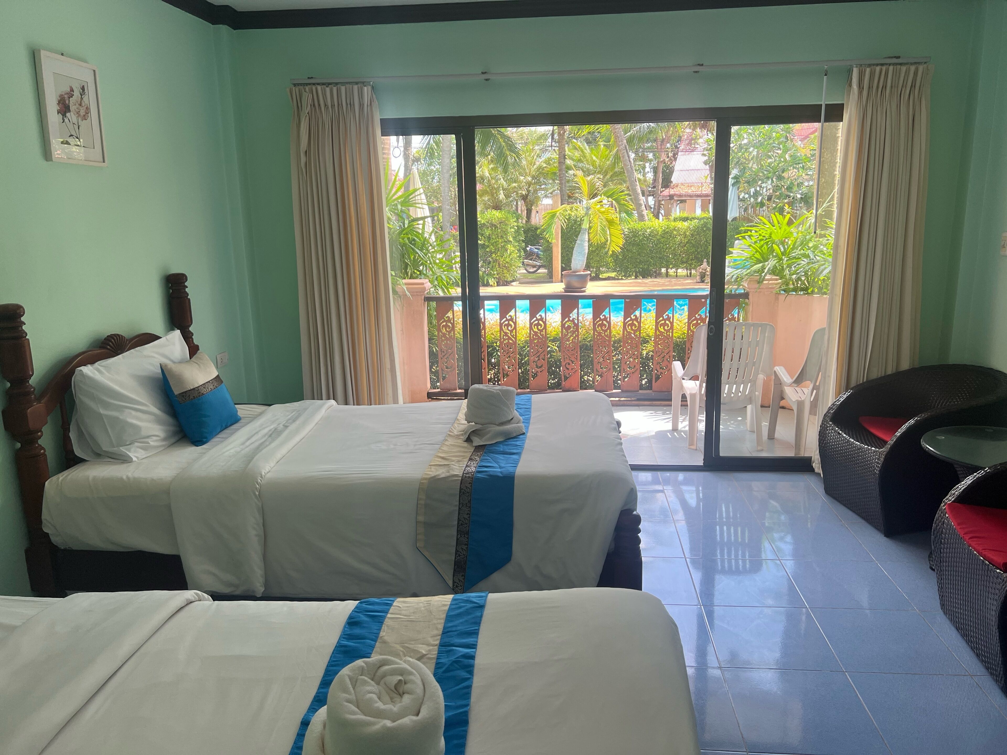 A hotel room with two beds, a balcony with a view of greenery, and a wicker chair.