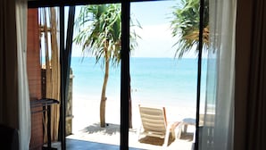 Vista da praia através de uma porta de correr de vidro, com palmeiras, uma cadeira de praia e uma mesa pequena.