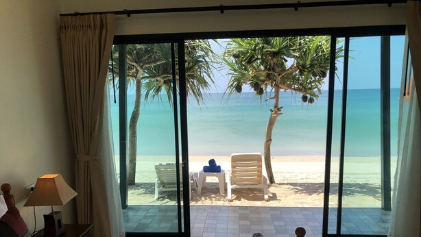 Quarto de hotel com uma grande janela com vista para uma praia, uma cama com edredom branco e um arranjo floral, e um abajur em uma mesa de cabeceira.