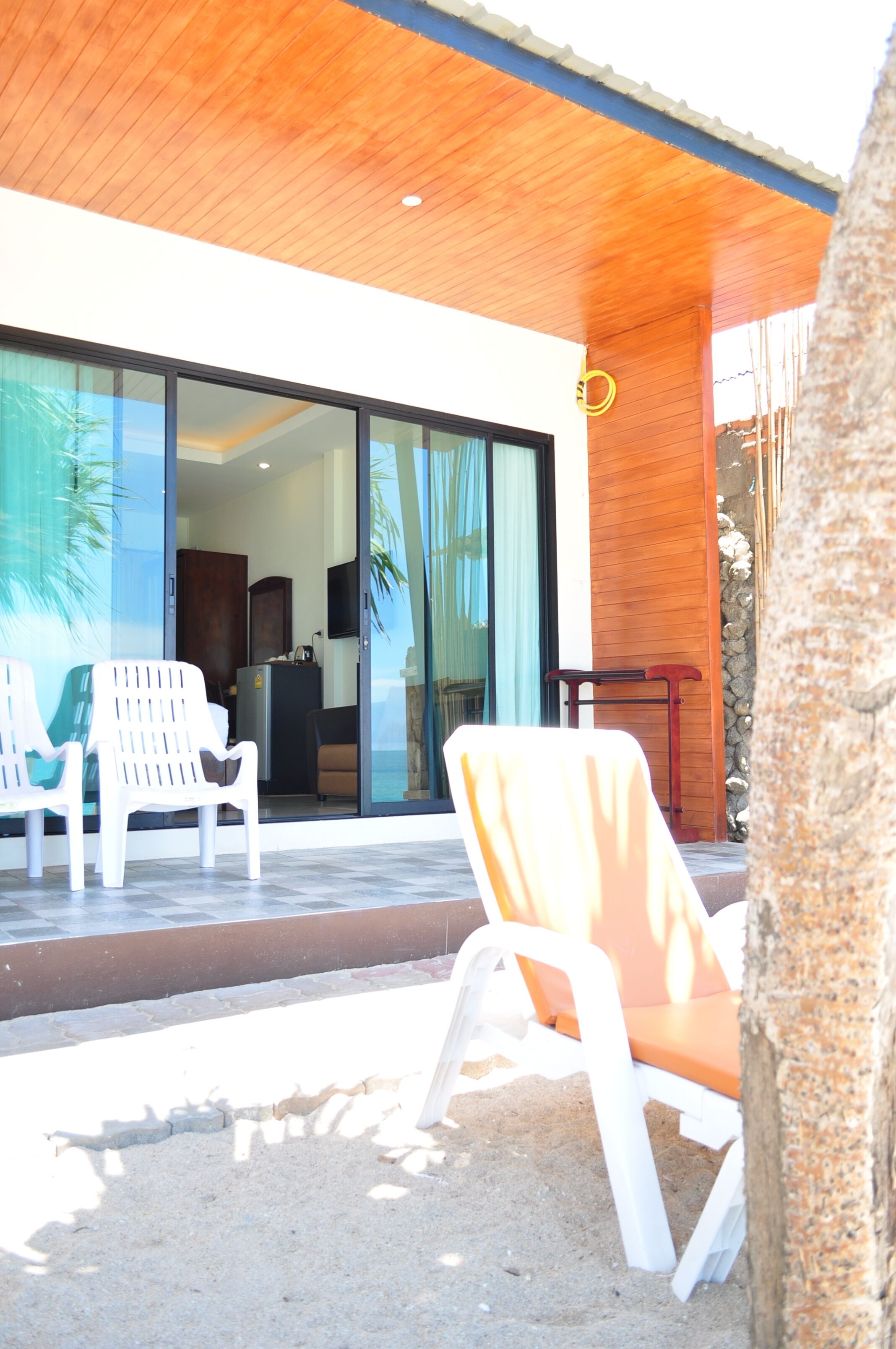 Un edificio moderno con techo de madera, puertas de cristal amplias y mobiliario exterior con sillas blancas y anaranjadas.
