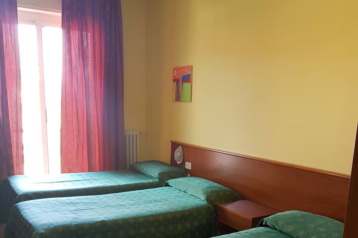 A room with two single beds, a wooden headboard, a radiator, and a window with red curtains.