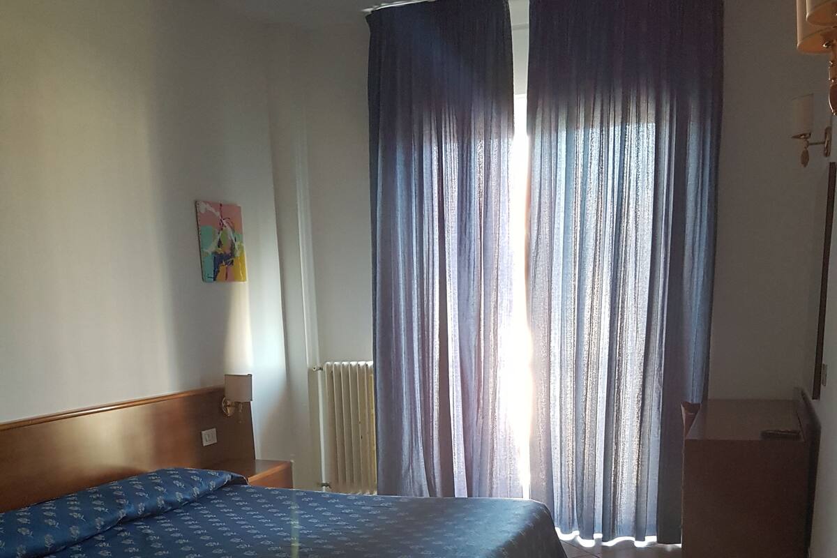 A hotel room with a bed, a radiator, a nightstand, a desk, and a window with curtains.