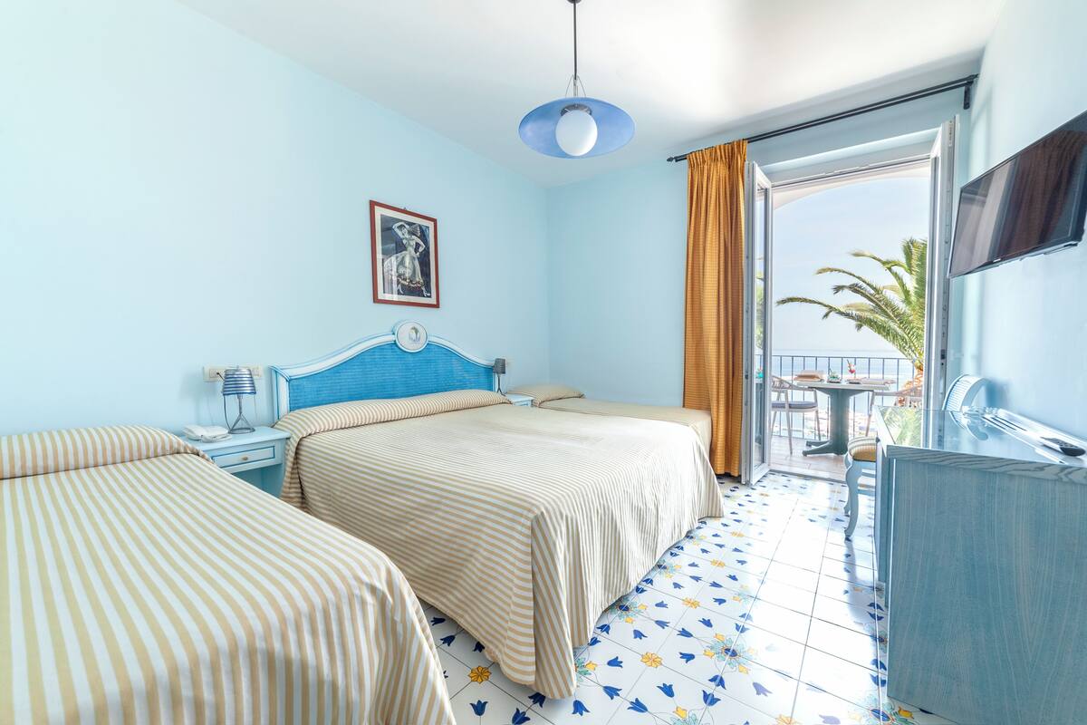 A hotel room with two beds, a desk, a chair, a TV, and a balcony with a view of palm trees.