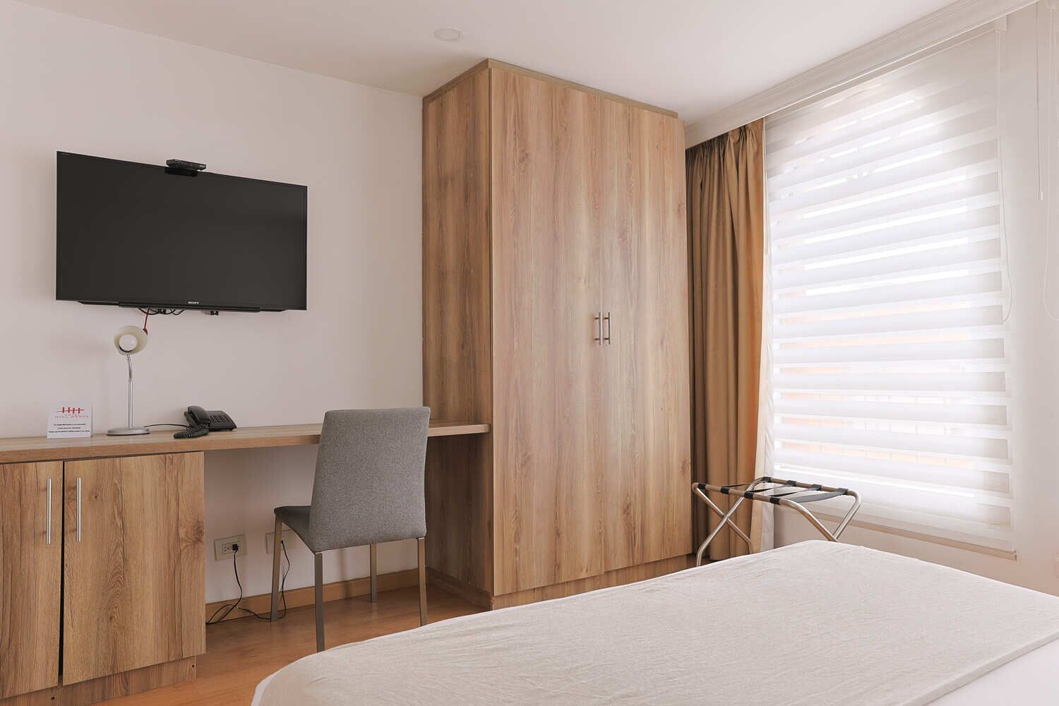 A hotel room with a bed, a desk with a chair, a TV mounted on the wall, and a window with blinds.