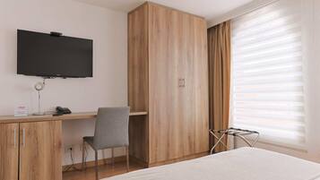 A hotel room with a bed, a desk with a chair, a TV mounted on the wall, and a window with blinds.