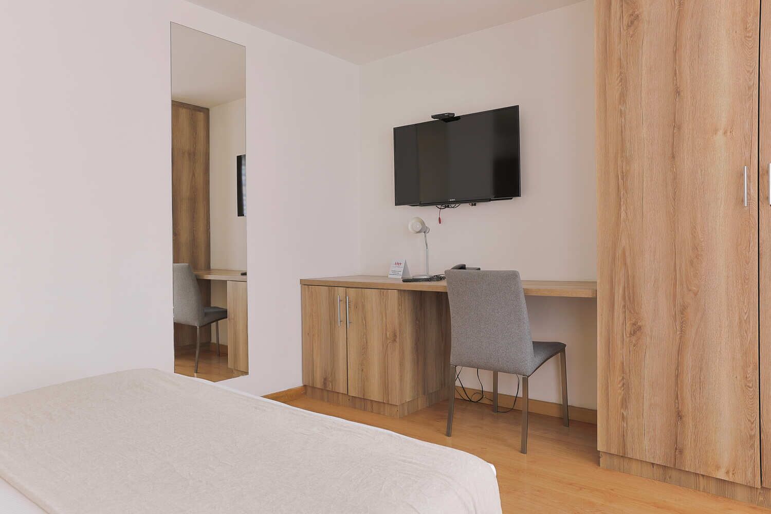 A bedroom with a bed, a desk with a chair, a TV mounted on the wall, and wooden wardrobe.