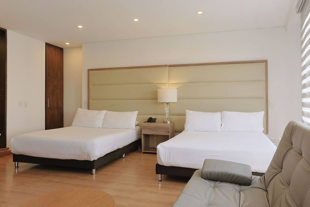 A hotel room with two beds, a nightstand, a lamp, and a sofa.