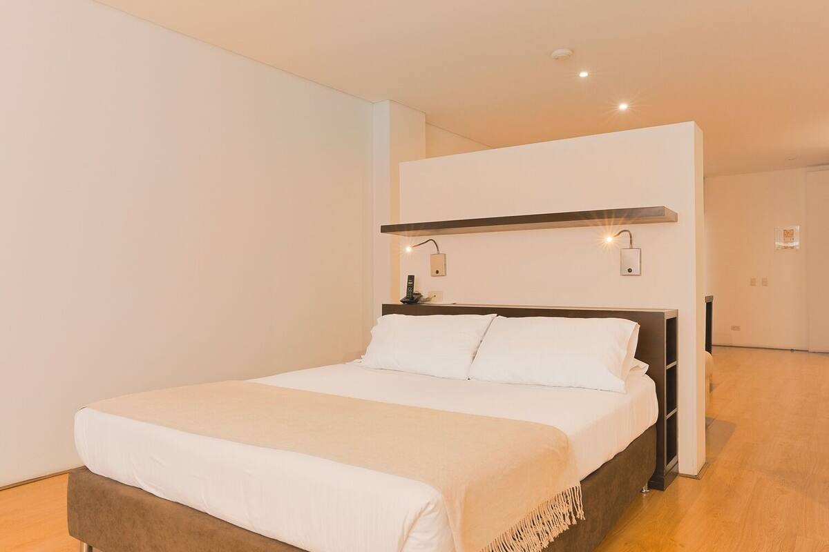 A modern hotel room with a large bed, a bedside table, and a wall-mounted light fixture.