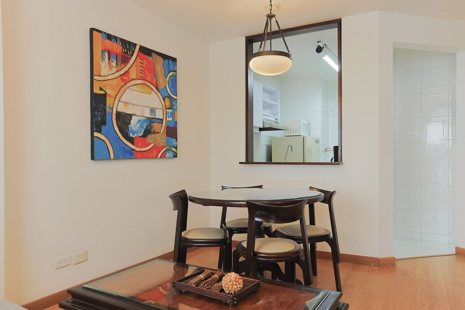 A dining area with a round table, four chairs, a hanging light fixture, and a colorful abstract painting on the wall.