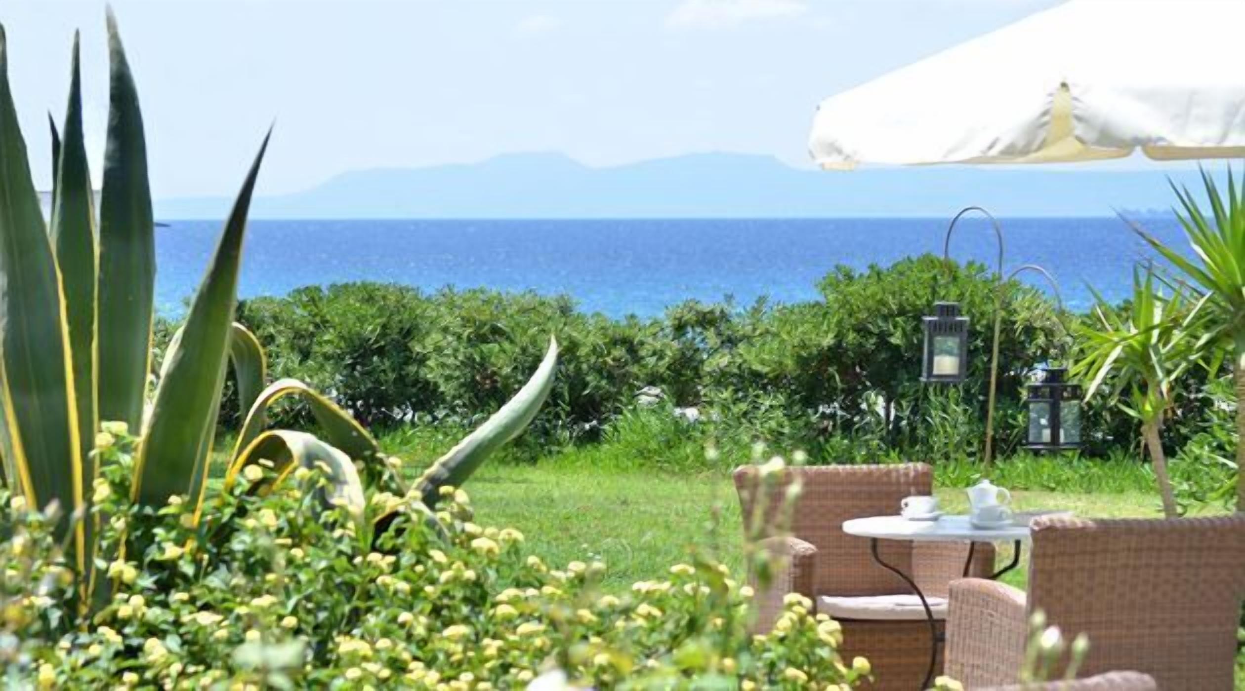 An outdoor seating area with a view of the sea, surrounded by greenery and a white umbrella.