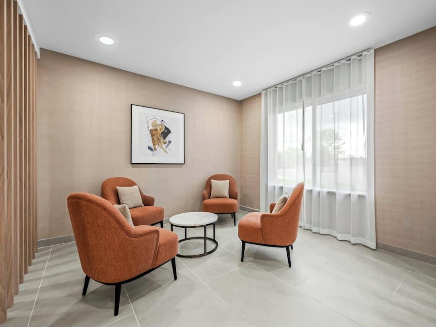 Quarto moderno com quatro poltronas laranja, uma mesa de centro redonda e uma janela grande com cortinas brancas.