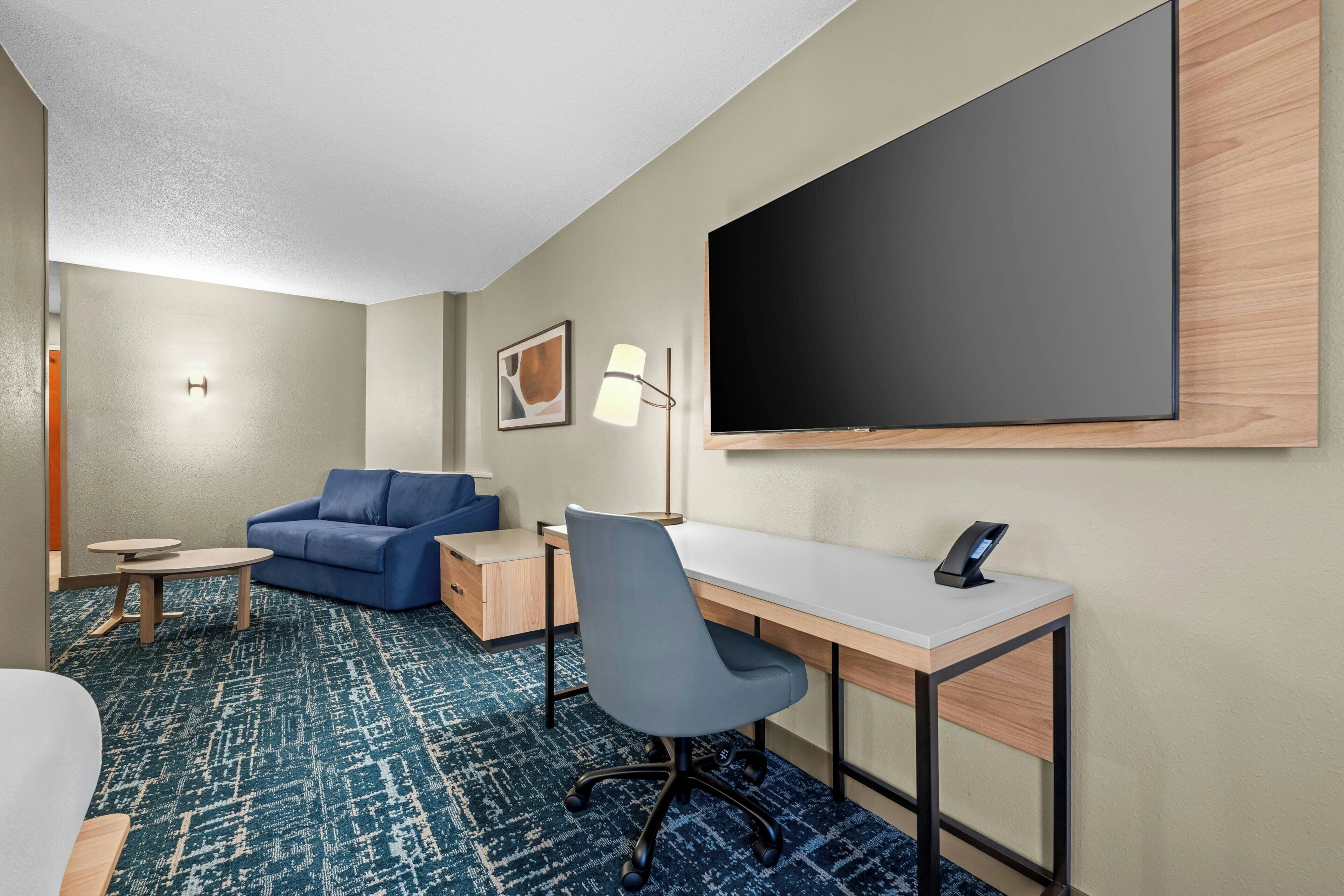 Habitación de hotel con cama, escritorio, silla, sofá, mesita pequeña, lámpara y televisor grande.