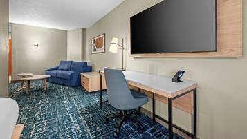 Habitación de hotel con cama, escritorio, silla, sofá, mesita pequeña, lámpara y televisor grande.