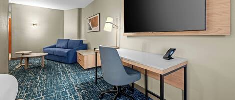 A hotel room with a bed, a desk, a chair, a sofa, a small table, a lamp, and a large TV.