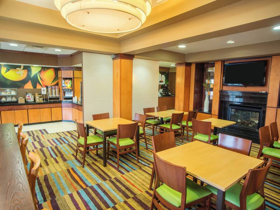 A hotel dining area with multiple tables and chairs, a kitchen counter, and a television mounted on the wall.