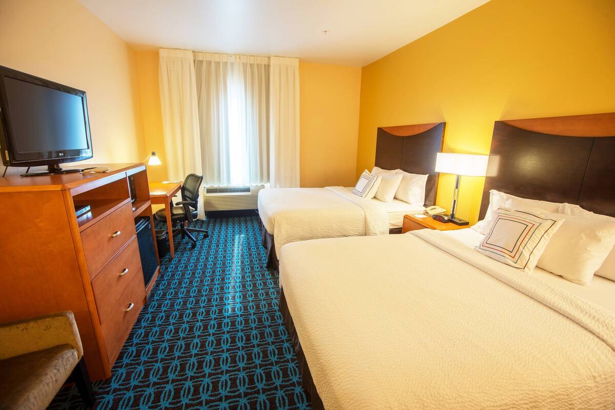 Ein Hotelzimmer mit zwei Betten, einem Schreibtisch mit Fernseher, einem Sessel und einem Fenster mit Vorhängen.