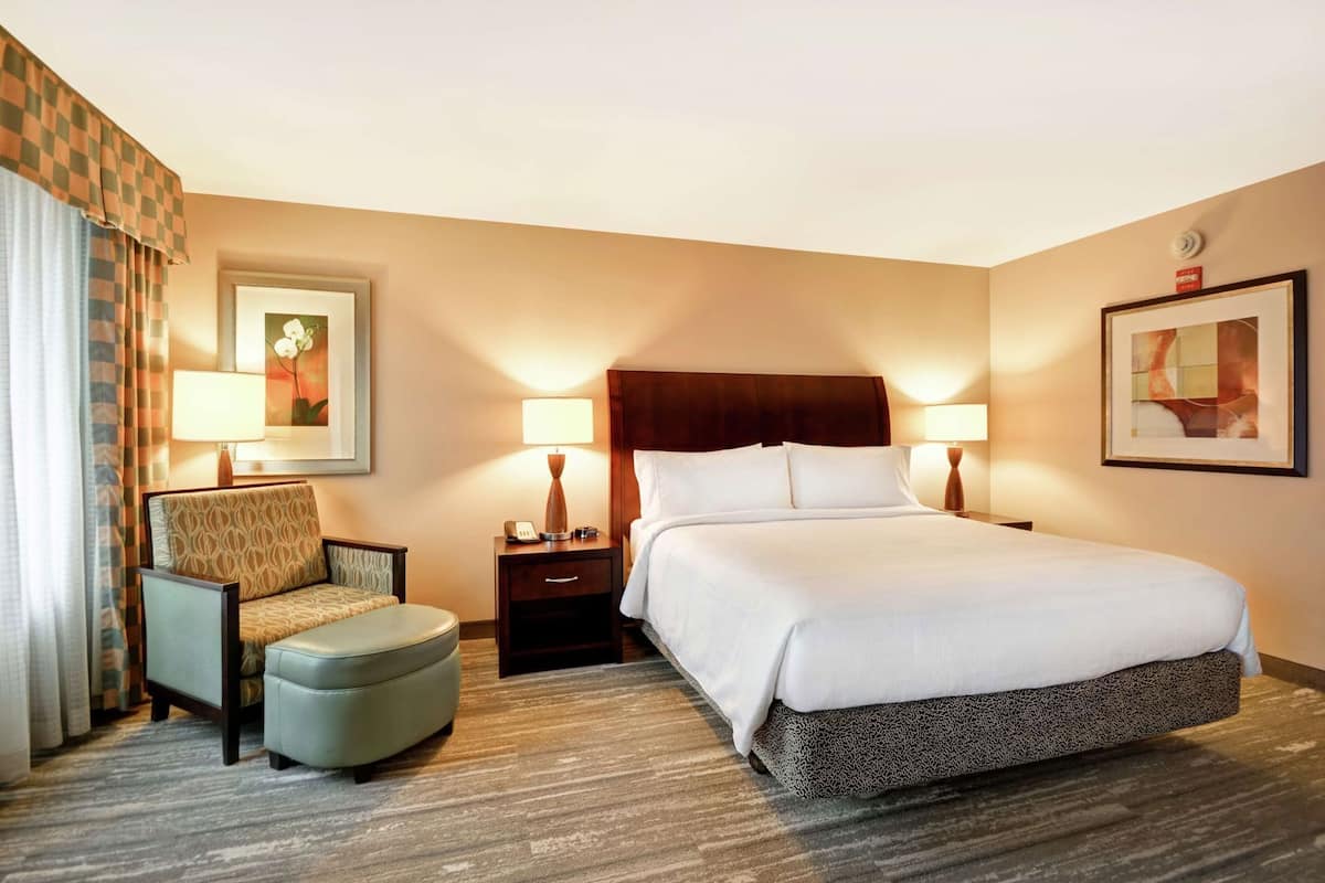 A hotel room with a bed, a chair, a nightstand, a lamp, and a framed picture on the wall.