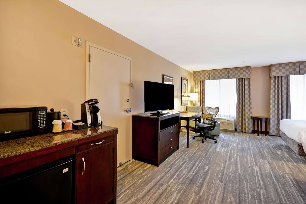 A hotel room with a kitchenette, a desk, a chair, a bed, and a window with curtains.