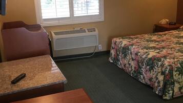 A hotel room with a bed, a desk, a chair, a window with blinds, a wall-mounted TV, and a framed picture.