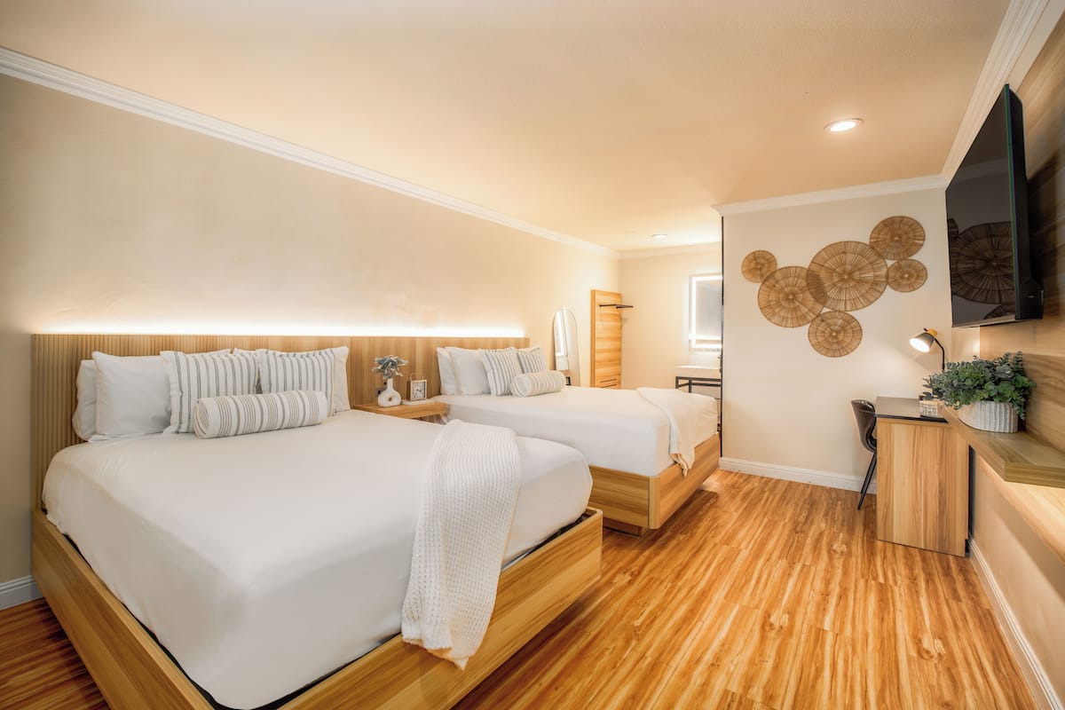 A hotel room with two beds, a wooden floor, a desk, and a wall-mounted TV.