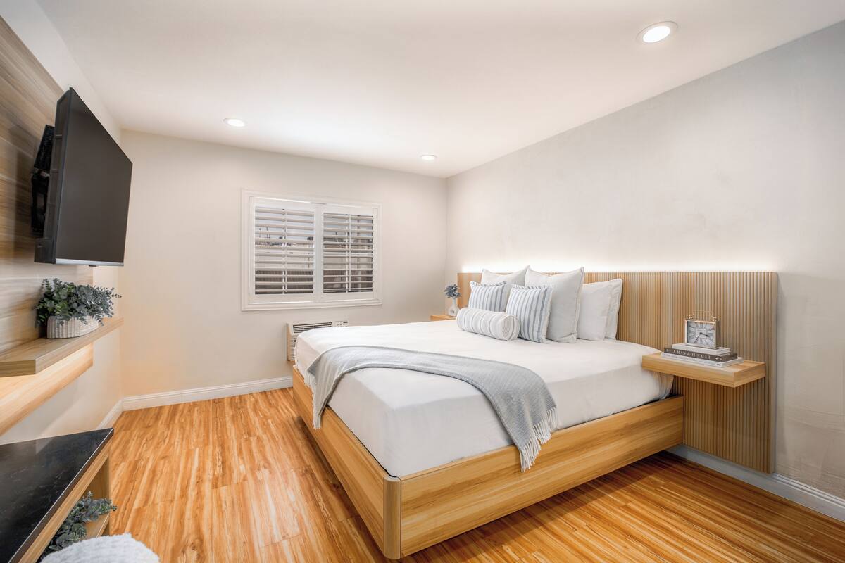A bedroom with a bed, bedside table, TV, window, and wooden flooring.