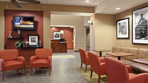Vestíbulo de hotel con sillones tapizados de color rojo, un televisor mostrando el pronóstico del tiempo, cuadros enmarcados y una mesa de recepción.