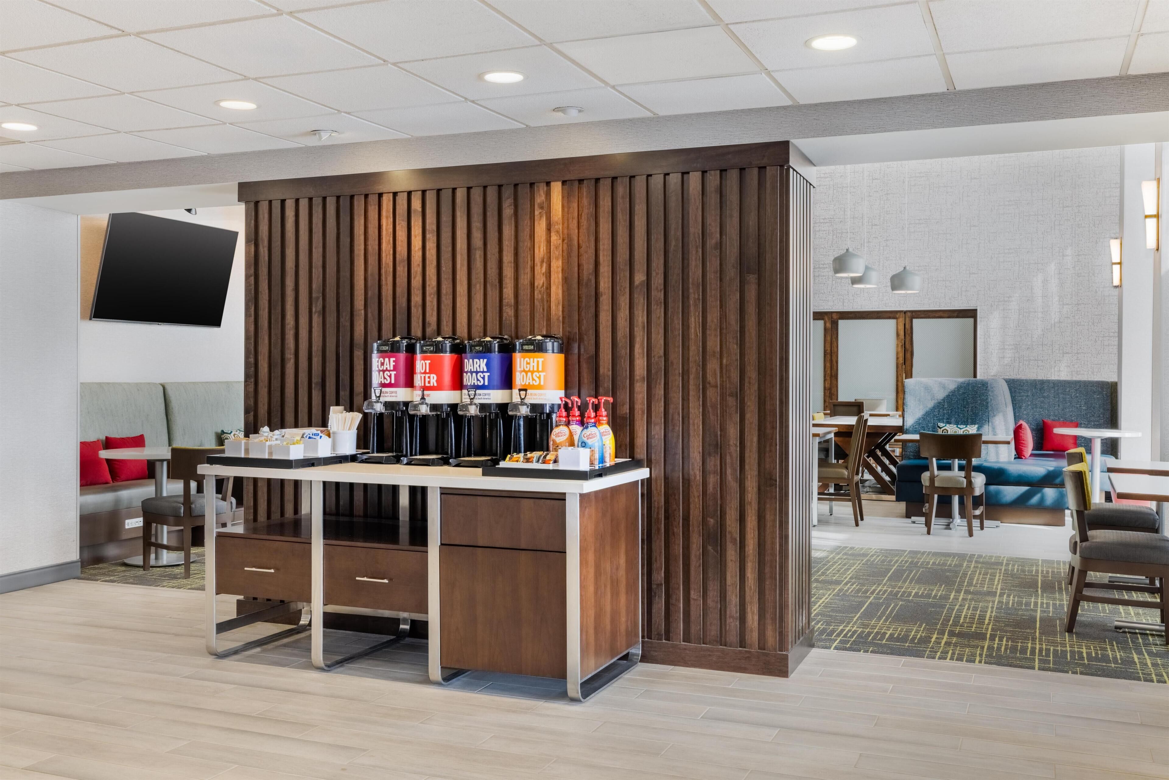 A modern hotel lobby with a coffee station, seating area, and a TV mounted on the wall.
