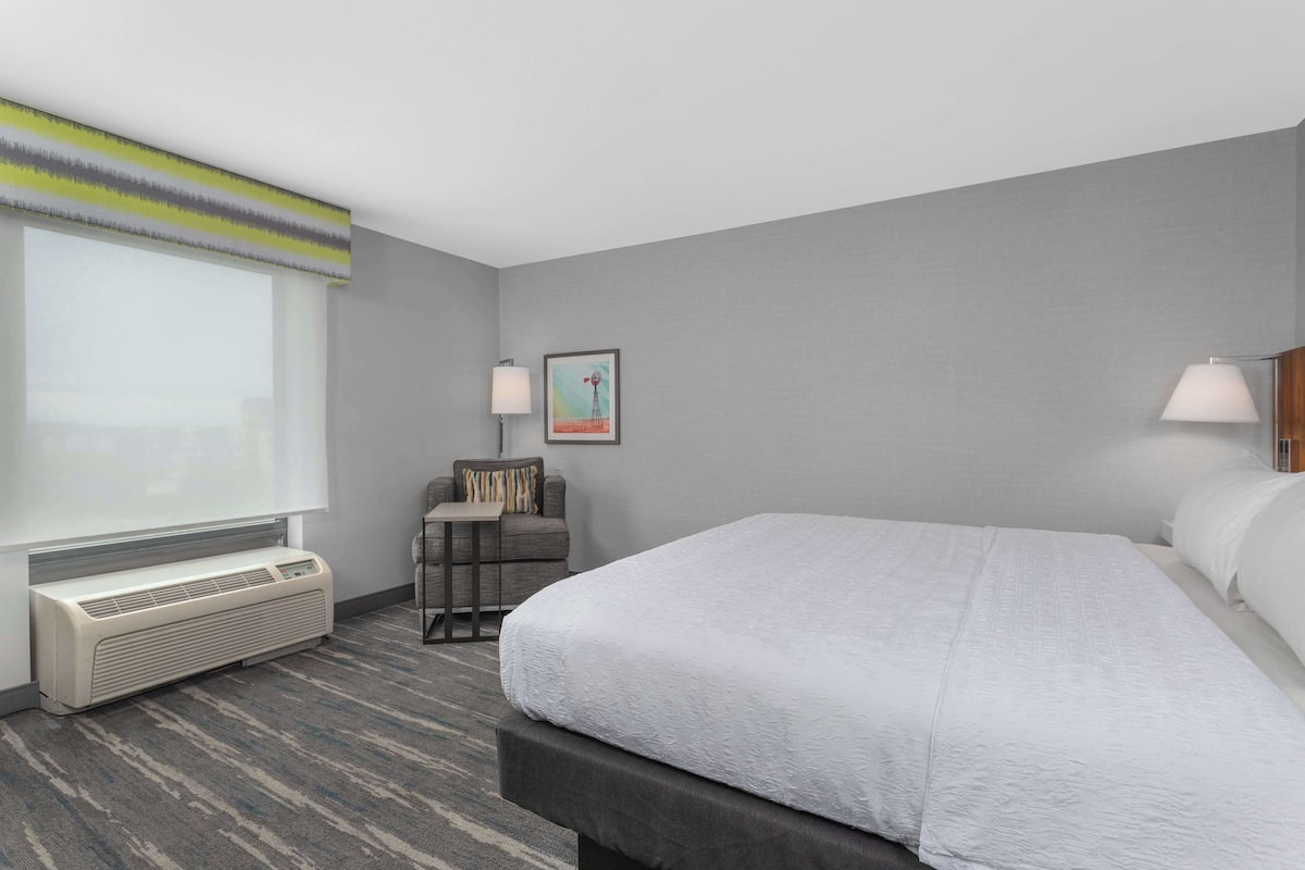 A hotel room with a large bed, a desk, a chair, a window, and a wall-mounted air conditioner.