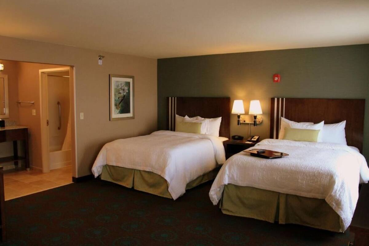 A hotel room with two beds, a desk, a chair, a lamp, and a painting on the wall.