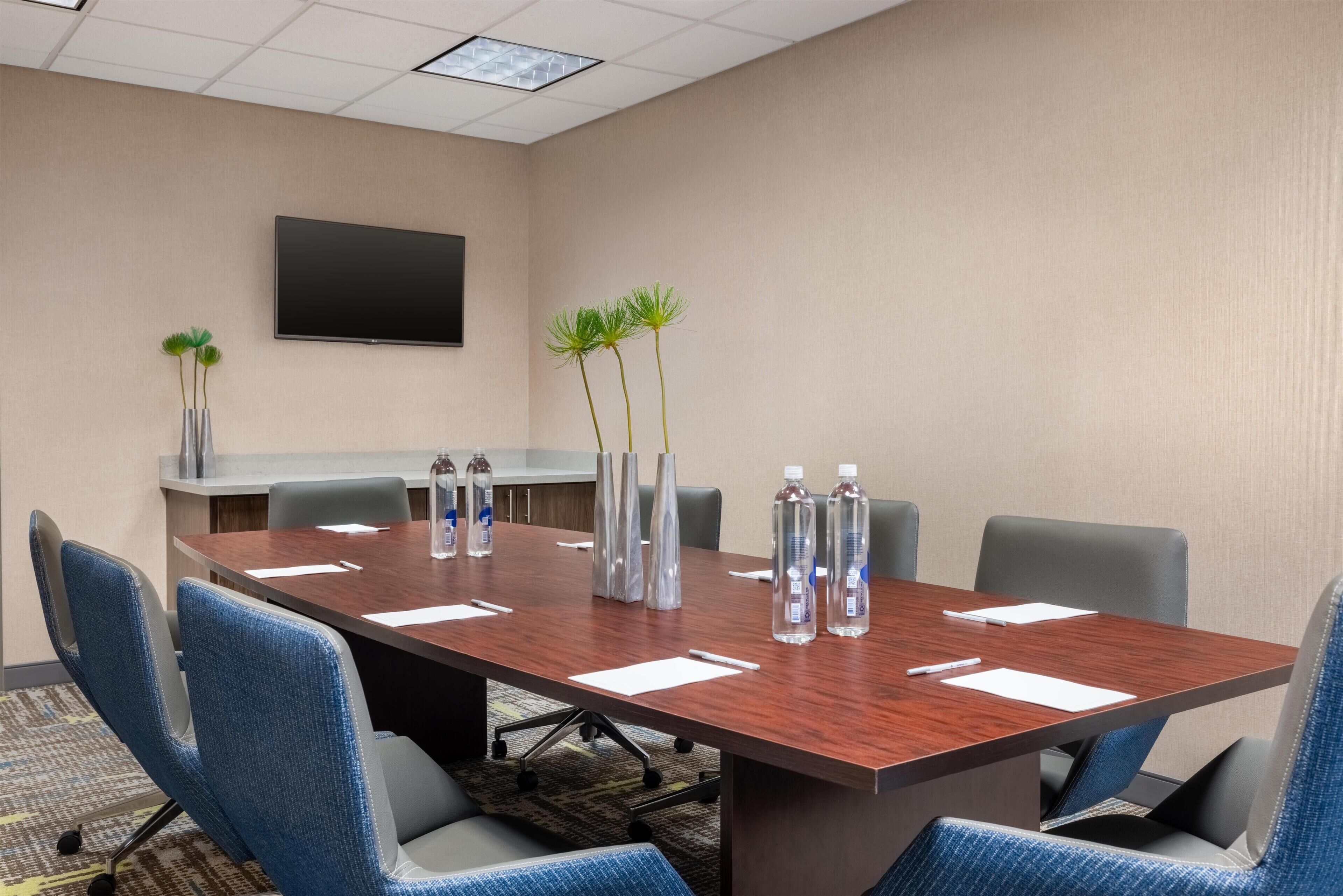 Un salón de conferencias con una larga mesa, sillas, botellas de agua y un televisor en la pared.